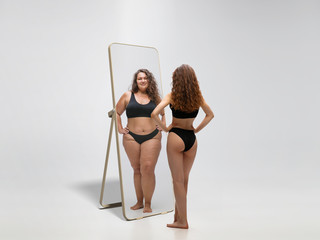 Young fit, slim woman looking at fat girl in mirror's reflection on white background. Thinking she's not enough sportive. Concept of healthy lifestyle, fitness, sport, nutrition and body positive.