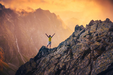 Happy young man in mountains. Positive mind in beautiful nature. Alone man hold hands in air, beautiful sunset hilly landscape in background. Orange and yellow edit space..