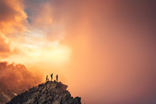 Together Overcoming Obstacles As A Group Of Three People Up On The Top Of A Mountain. Celebrate Victory And Success Over Sunset Background. Goal Achievement Symbol..