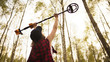 Treasure hunter, young woman holding metal detector in the air in the forest. Low angle. High quality photo