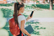 © Maridav - Back to school semester online on phone app. University student using mobile phone for watching her classes while walking on campus with backpack, laptop, book. Woman holding cellphone.