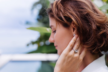 Fashion portrait of woman at tropical luxury villa wearing white stylish blazer, rings, earings on background of tropical leaves and swimming pool.