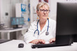 © DC Studio - Mature doctor typing patient report on computer in hospital cabinet wearing stethoscope around the neck