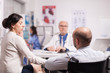 © DC Studio - Disabled young man in wheelchair and his wife during consultation with senior doctor in hospital office. Elderly aged doctor wearing white coat and stethoscope. Nurse with blue uniform in the