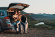 © Romvy - Young Traveler Couple on a Road Trip, Man and Woman Sitting on the Opened Trunk of Their Car Over Sunset