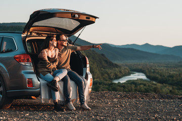 young traveler couple on a road trip, man and woman sitting on the opened trunk of their car over su