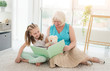© Ievgen Skrypko - Cute granny reading book to little granddaughter holding toy sitting on floor in playroom