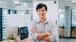 © tirachard - Portrait of successful handsome executive businessman smart casual wear looking at camera and smiling, arms crossed in modern office workplace. Young Asia guy standing in contemporary meeting room.