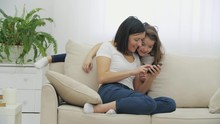 Young Mother Sitting On The Sofa With Phone In Hands, Looking Over The Photos. Beautiful Daughter With Long Hair Is Leaning Over The Back Of The Sofa, Also Looking At The Screen.
