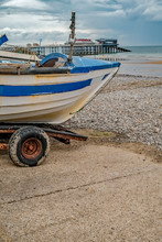 A Close Up Of A Traditional Crab Fishing Boat Moored On The Shingle Beach In Cromer After A Morning At Sea. In The Distance Is The Victorian Pier.