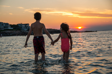 Wall Mural - Brother and sister holding hands in the ocean at sunset