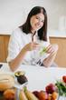 © sonyachny - Young happy woman holding green lettuce leaf in hands and smiling on background of fresh fruits and vegetables in modern white kitchen. Healthy food concept. Home cooking