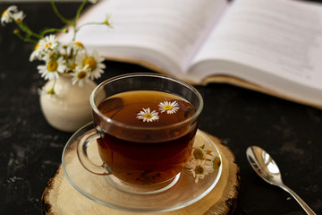 English tea with chamomile stands on a wooden frame and a teaspoon and an open book for reading lies on a black background. Nearby is a white vase with summer chamomile flowers