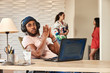 © SuperStock - portrait young ethnic man sitting at desk with laptop computer wearing large over ear headphones, coworkers standing in background talking