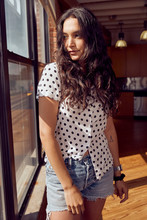 Portrait Of Young Ethnic Woman Wearing Polka Dot Blouse And Cut Off Jean Shorts Standing By Bank Of Windows In Downtown Loft 