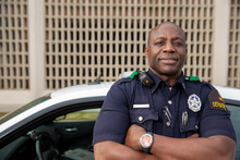 Portrait Of Police Officer Standing In Street In Front Of Squad Car Looking Towards Camera With Arms Behind His Back 