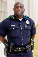 Portrait Of Police Officer Standing In Street In Front Of Squad Car Looking Towards Camera With Arms Behind His Back 