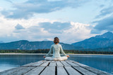 Fototapeta  - Mid adult woman meditating while sitting on jetty over lake against cloudy sky