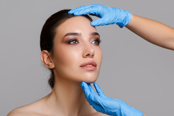 Facial beauty. Portrait of a beautiful young woman with perfect makeup and soft smooth skin. Close-up of beautician hands in gloves touching the face. Plastic surgery concept.