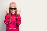 Fototapeta Do pokoju - Young woman in a hat and glasses and a pink sports jacket makes a rock and roll goat gesture, we'll catch this party on a white background with empty space. Concept. Face expression. Banner