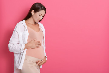 Wall Mural - Beautiful pregnant woman hugging her tummy against pink background, looking at her belly with love, copy space for advertisement or promotional text text.