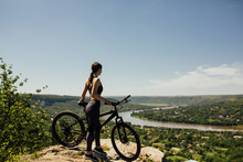 Woman Cyclist Standing With Bicycle On Rock On Mountain. Young Athlete Biker With Mountain Bike On Top Of The Hill Observing The River And Town View Under Blue Sky.