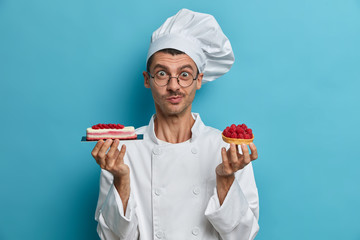 Cooking, profession, bakery concept. Young male cooker holds tasty confectionery, desserts decorated with berries, poses at restaurant kitchen, isolated over blue background, has creative occupation