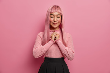 Wall Mural - Studio shot of dreamy pleased Asian woman stands with eyes closed, smiles happily, has long pink hair, wears sweater, black skirt, poses against rosy background. Positive emotions and feelings.