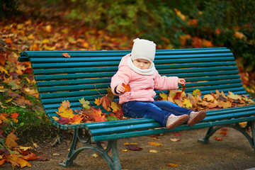 Wall Mural - Adorable toddler girl sitting on the bench and playing with fallen leaves