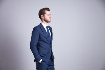 A businessman in a blue business office suit holds his hands in his pockets and looks to the side with a serious face.