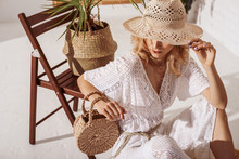Elegant Fashionable Woman Wearing Summer White Crochet Jumpsuit, Hiding Her Face With Straw Hat, Holding Wicker Bag, Posing In Stylish Boho Interior. Copy, Empty Space For Text