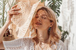 © Victoria Fox - Happy smiling woman enjoying sunlight, hiding her face with straw hat, wearing trendy necklace with many chains, posing at home, in stylish boho interior. Summer fashion conception