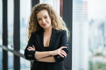 Young beautiful business woman standing arms crossed near window in office. Smiling sexy lady confident in black suited.