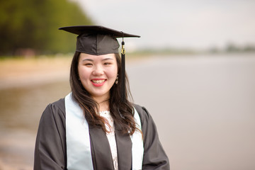 Happy Asian student holding degree certificate file while wearing gown and mortarboard in the outdoor.Young female Chinese student graduate with graduation hat.