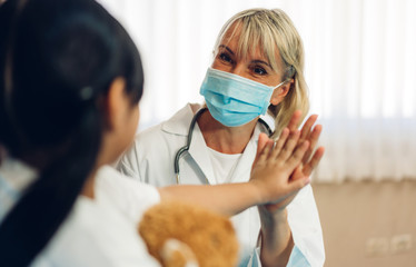 Woman doctor wearing protective mask service help support discussing and consulting talk to little girl patient and giving hi five check up information in hospital