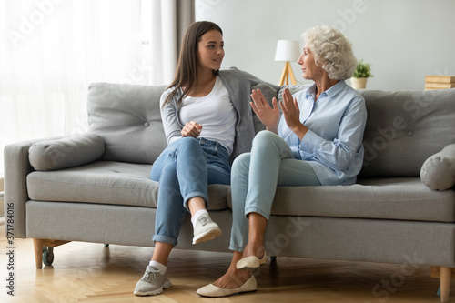 Full length young happy beautiful woman sitting on comfortable couch with smiling middle aged 60s mother, enjoying pleasant conversation in living room, discussing sharing life moments indoors.
