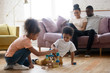 © fizkes - Calm bliss. Two little african kids sister and brother playing on wooden heated floor at living room building toy house from blocks and parents mum and dad sitting on sofa, hugging and using tablet pc