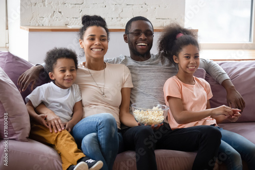 Lazy weekend. Happy smiling african family millennial married couple with two kids elder daughter and little son embracing, sitting on cozy sofa and eating popcorn before tv screen watching nice movie