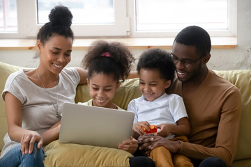 Wall Mural - Happy laughing african family, young dad and mom with two little kids sister and brother sitting on cozy couch at lounge using laptop, playing online game by wifi internet, shopping in virtual store