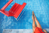 Fototapeta Przestrzenne - Cropped top view of sexy woman feet soaking in swimming pool. Attractive female legs in bikini, enjoying cool water at poolside, sit on pool edge near red lifeguard chair, travel and tourism concept