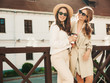 © halayalex - Two young beautiful smiling hipster girls in trendy white sweater and coat. Sexy carefree women posing on street background in hat and sunglasses. Positive models having fun and drinking coffee