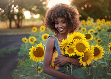 Happy Young Black Woman Walks In The Sunflower Field. Smiling Dark-skinned Girl With With A Bouquet Of Sunflowers And Curly Hair.