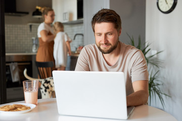 confident caucasian male work from home, sit in the kitchen with laptop, while his wife and daughter doing household chores in the background