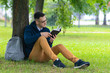 © Евгений Шемякин - Young concentrated guy in glasses, student reading an interesting book, leaning at a tree, relaxing in summer park with backpack. Handsome smart intelligent man studying outdoors.