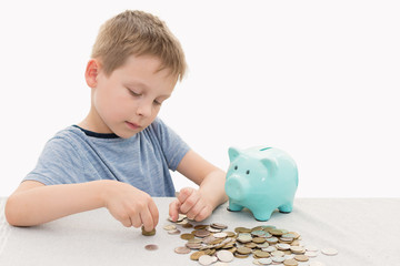 preschooler in a blue t-shirt on a white background. The child counts, examines and puts the accumulated coins into a piggy bank. The idea is to teach children how to save and value money.