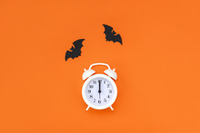 White Alarm Clock And Black Cardboard Bats On Orange Background. Halloween Minimal Concept. Top View, Copy Space