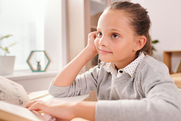 Smiling clever schoolgirl with open book looking through questions on blackboard