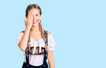 Beautiful caucasian woman with blonde hair wearing octoberfest traditional clothes covering one eye with hand, confident smile on face and surprise emotion.