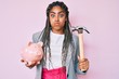 © Krakenimages.com - Young african american woman with braids holding piggy bank and hammer puffing cheeks with funny face. mouth inflated with air, catching air.