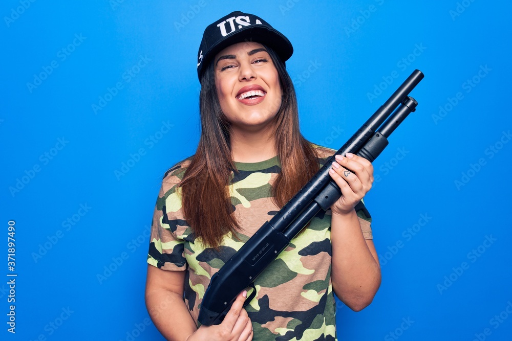 Young beautiful hunter woman wearing camouflage t-shirt and usa cap holding shotgun looking positive and happy standing and smiling with a confident smile showing teeth